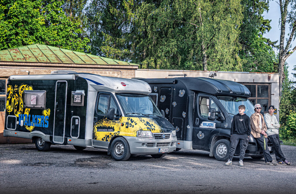 Två husbilar står parkerade. Framför dem står tre unga män.