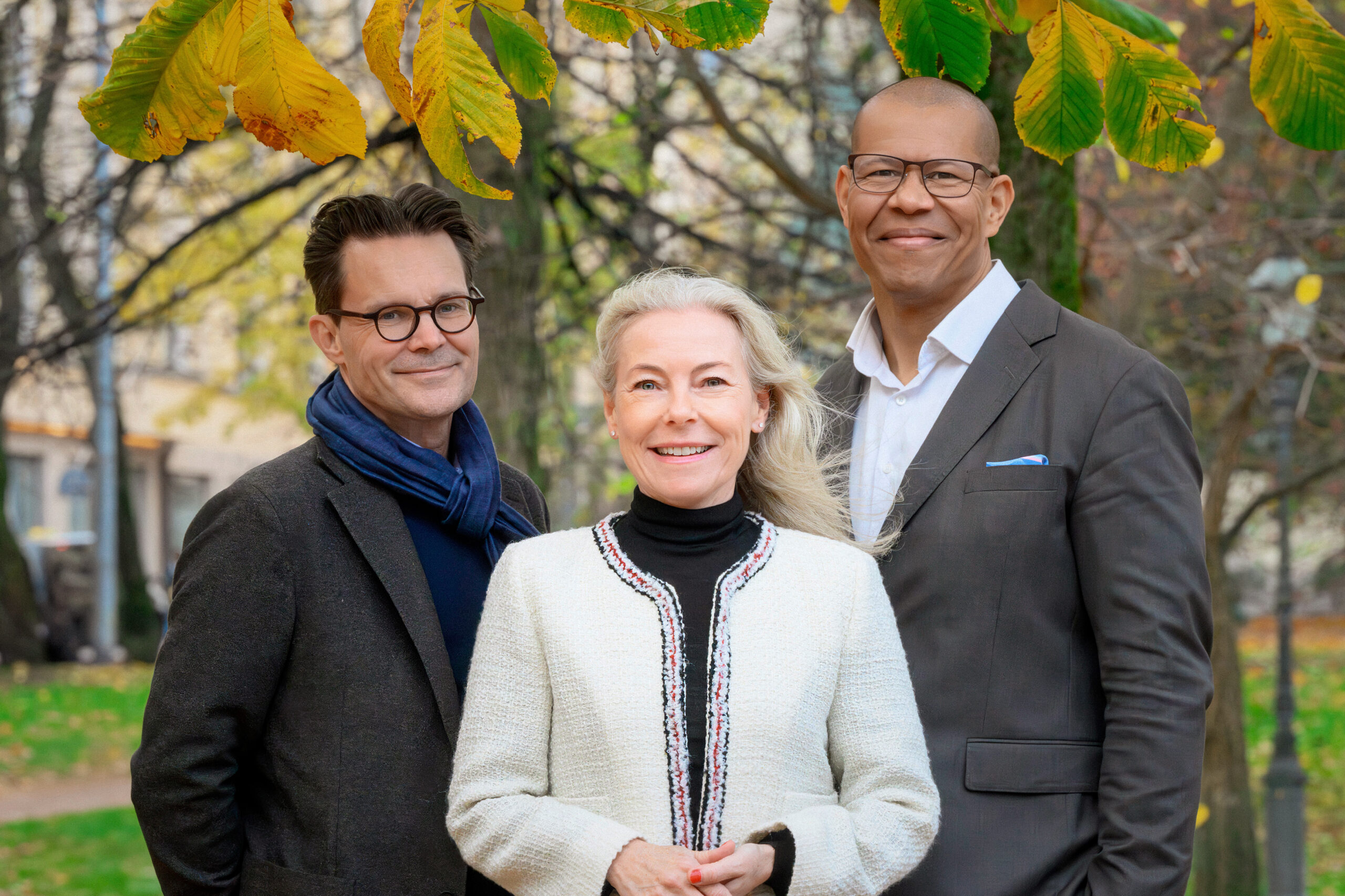 Tre personer, två män och en kvinna ler mot kameran. De är fotade utomhus där träden i bakgrunden har höstfärger.