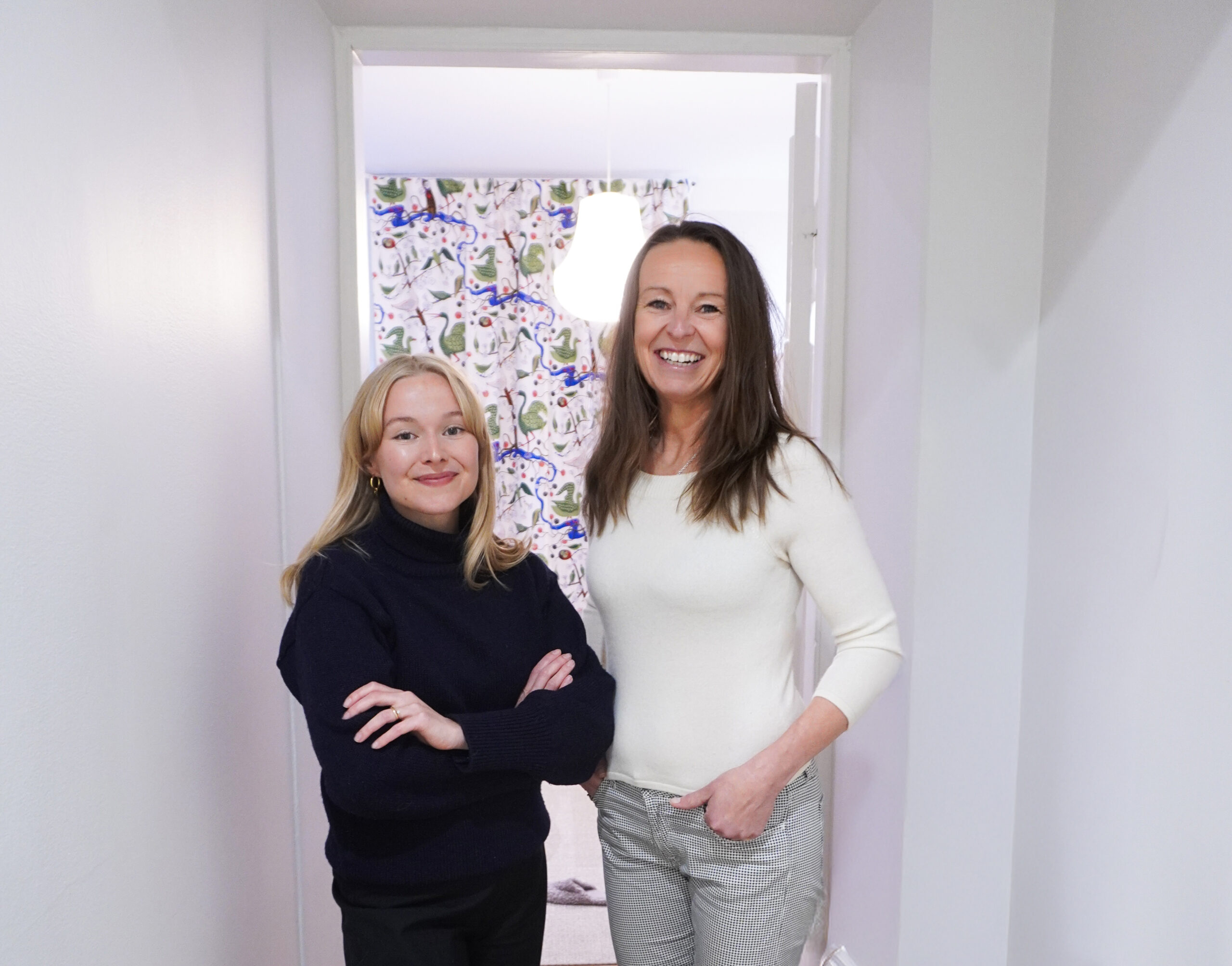 Två leende kvinnor tittar mot kameran. Bilden är tagen inomhus, i en ljus korridor.