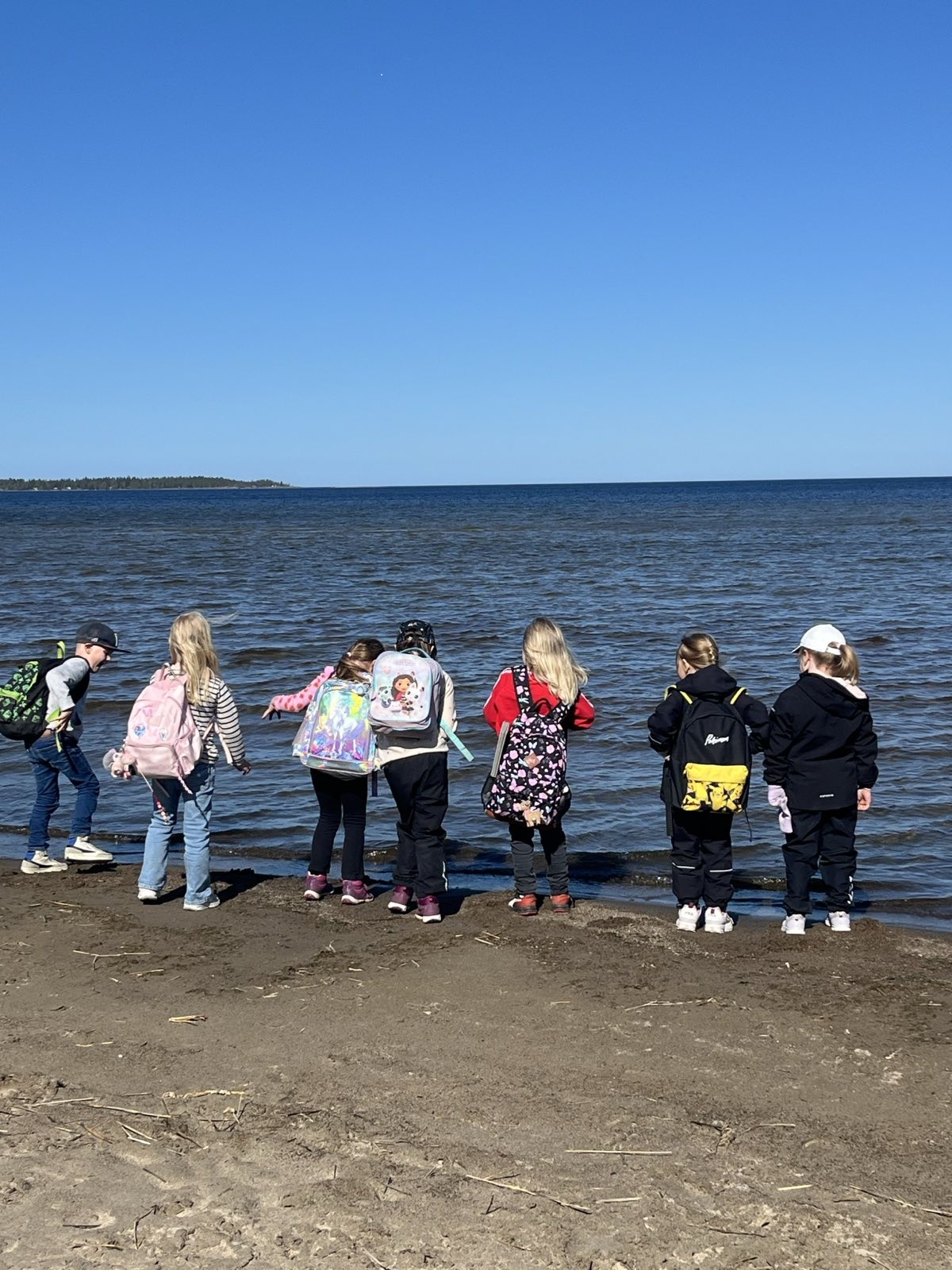 Sju skolbarn med ryggsäckar på ryggen står i vattenbrynet på en sandstrand med öppet hav i bakgrunden.