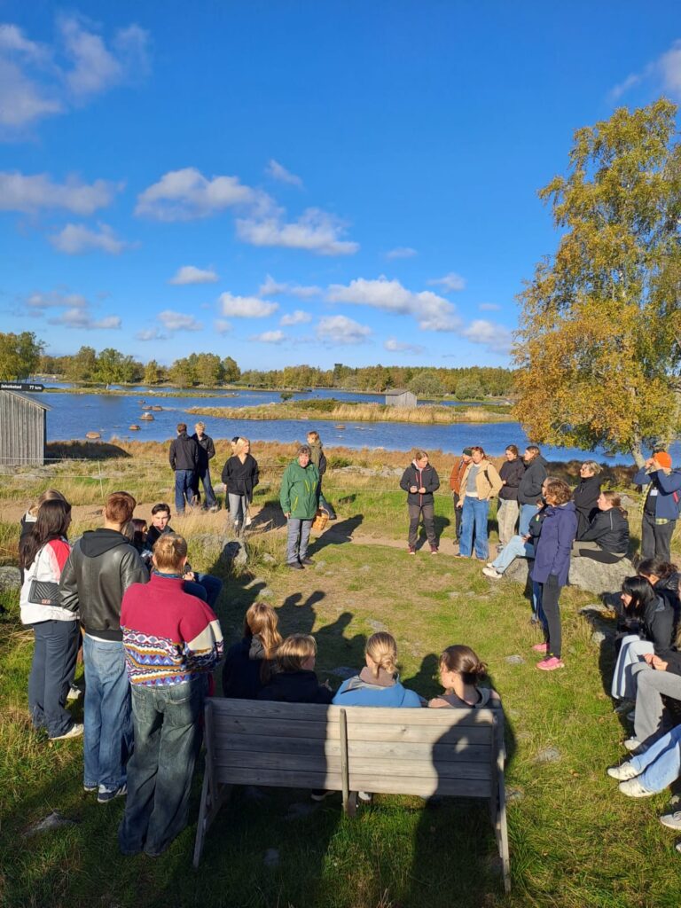 Skolelever utomhus i grön miljö med hav och skär i bakgrunden.